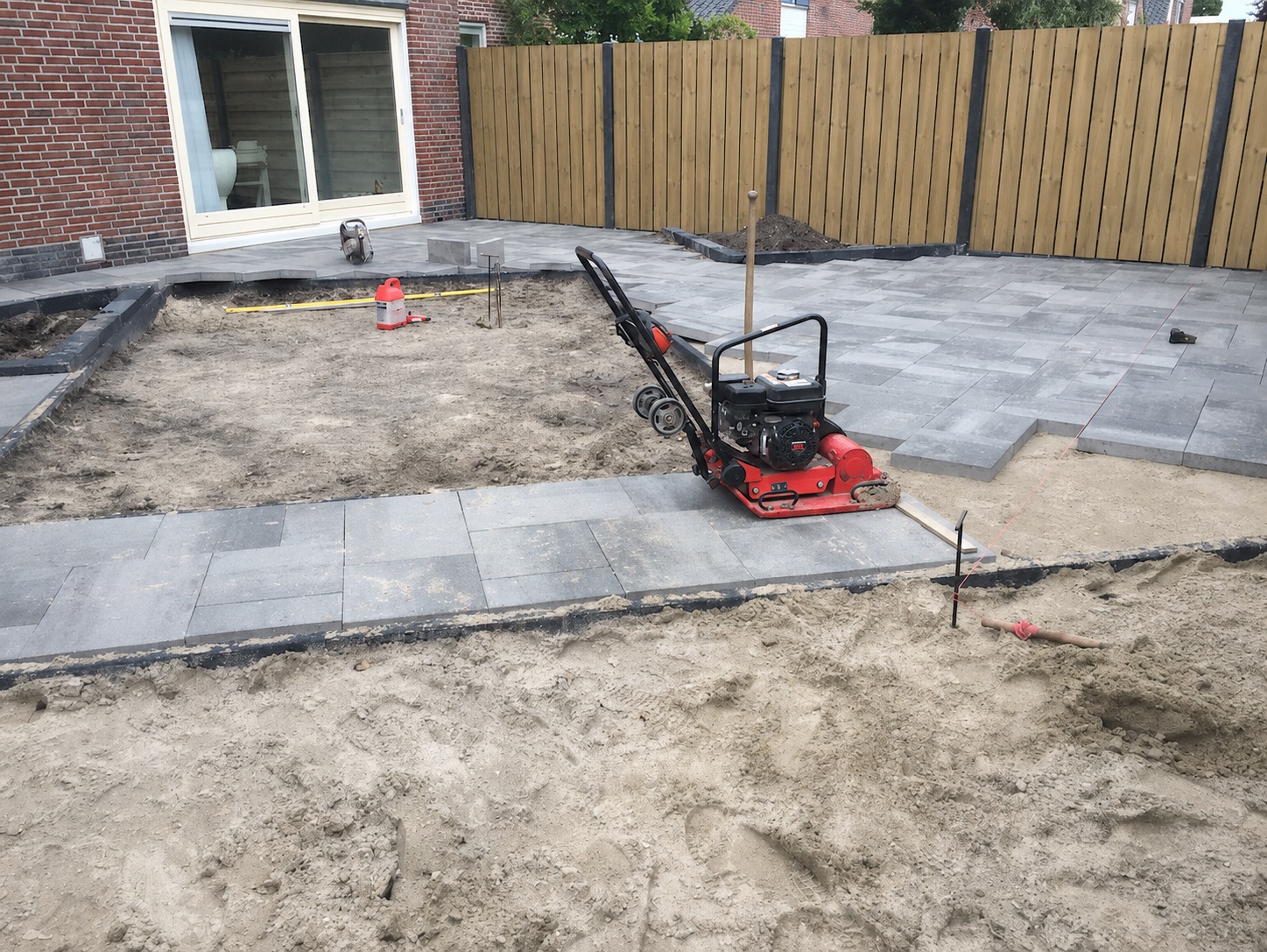 Tuinaanleg in uitvoering: grijze bestrating wordt op een zandbed gelegd en met een trilplaat verdicht, met houten schutting op de achtergrond