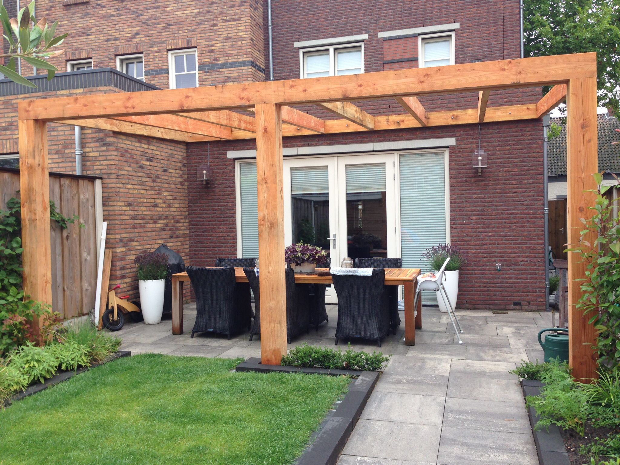 Houten pergola tegen de gevel boven een bestraat terras met eettafel en stoelen, met gazon op de voorgrond