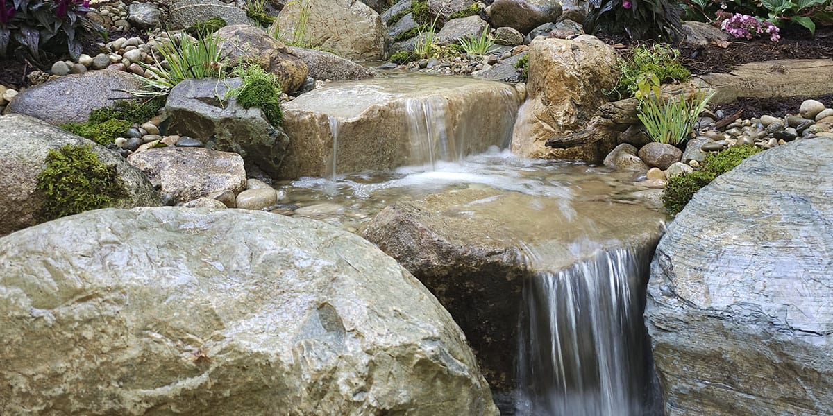 Natuurlijke miniwaterval van natuursteen met stromend water, omringd door grote keien, grind en mos